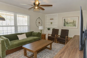 Ein Wohnzimmer mit einem grünen Sofa, einem Holztisch, zwei braunen Sesseln, einem Holzschrank, einer Uhr und einem Deckenventilator.