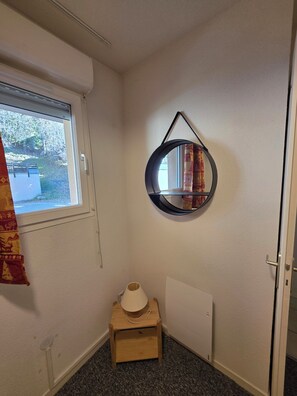 Ein Zimmer mit Fenster, einem runden Spiegel an der Wand, einem kleinen Holzschemel mit Lampe und einer Tür.