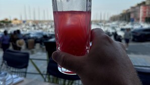 Una mano sosteniendo un vaso de bebida roja en un restaurante frente al mar, con mesas y sillas.