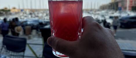 A hand holding a glass of red drink at a seaside restaurant with tables and chairs.