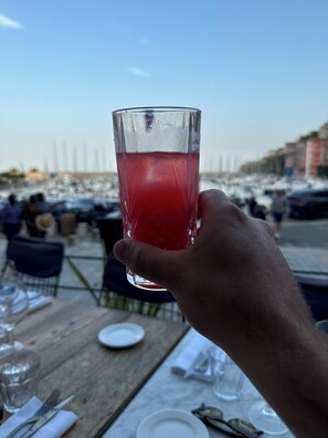 A hand holding a glass of red drink at a seaside restaurant with tables and chairs. - Village house (Murato)