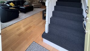 Una escalera que conduce a un piso superior, una sala de estar con un sofá y una mesa de centro, y una puerta al final de la escalera.