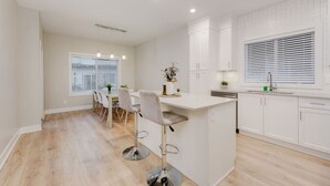 Cozinha moderna com armários brancos, ilha central com banquetas, área de refeições com mesa e cadeiras, e janela com persianas.
