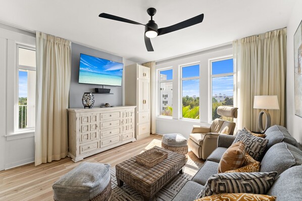 A spacious living room with a ceiling fan, large windows, a flat-screen TV, a white cabinet, a sofa, and a wicker ottoman. - Elegant Baytowne Wharf Luxury Getaway (Miramar Beach)
