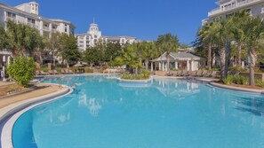 A large pool surrounded by palm trees and lounge chairs, with a building featuring a tower in the background. - Elegant Baytowne Wharf Luxury Getaway (Miramar Beach)