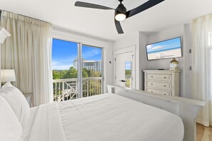 A bedroom with a bed, a ceiling fan, a TV, a dresser, and a balcony with a view of buildings and trees. - Elegant Baytowne Wharf Luxury Getaway (Miramar Beach)