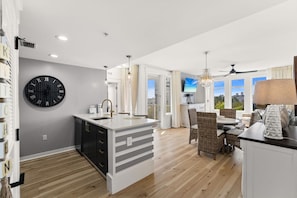A modern kitchen with a large clock, a dining area with wicker furniture, and a view of the outdoors. - Elegant Baytowne Wharf Luxury Getaway (Miramar Beach)