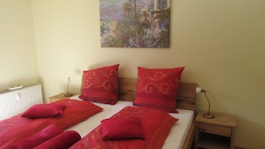 A neatly made bed with red bedding and pillows, a bedside table with a lamp, and a painting on the wall.