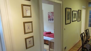 A hallway with framed pictures on the walls, a wooden floor, and a bed with a red pillow in the background.