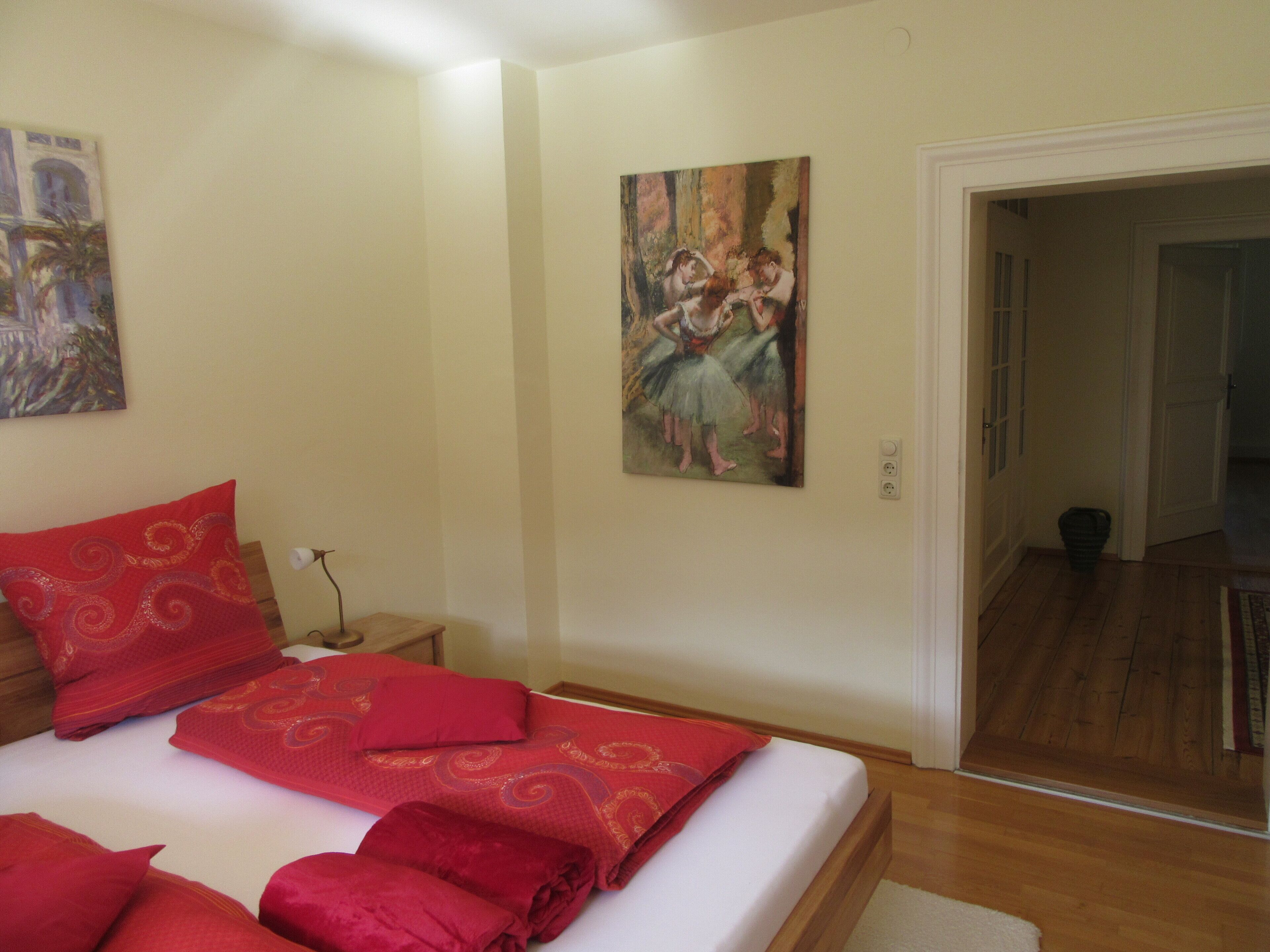 A bedroom with a bed, pillows, a nightstand, a painting, and a hallway leading to another room.
