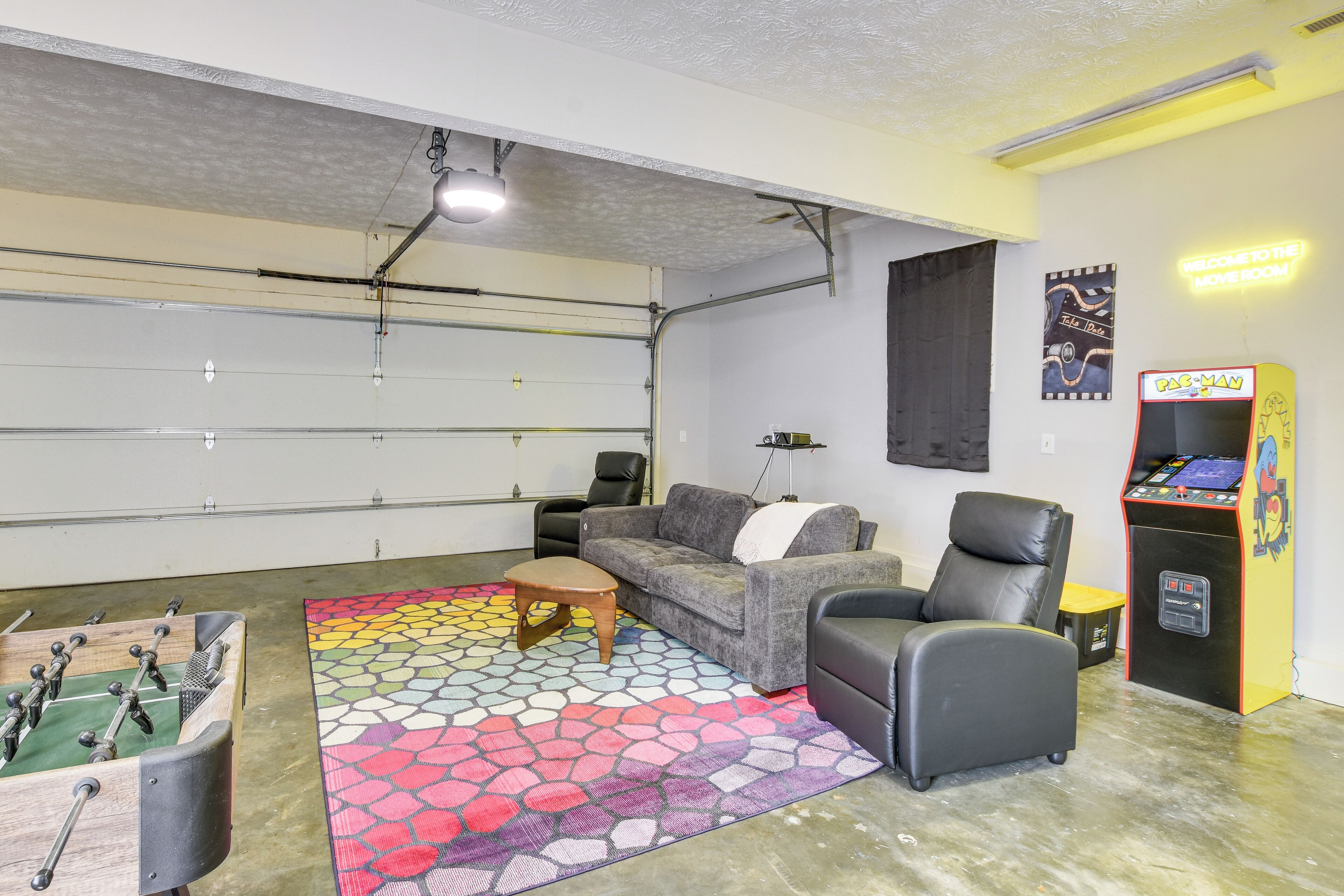 A garage with a foosball table, a colorful rug, a sofa, a chair, and a Pac-Man arcade machine.