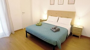 Een keurig opgemaakt bed met een blauwe sprei en kussens, een houten vloer, een witte deur en een nachtkastje met een lamp.