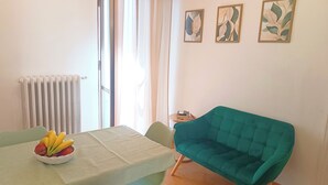 Een eetgedeelte met een tafel, stoelen, een schaal fruit, een radiator, een raam met gordijnen en kunst aan de muur.