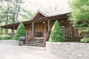 A wooden cabin with a porch, steps leading to the entrance, and two coniferous trees in front.