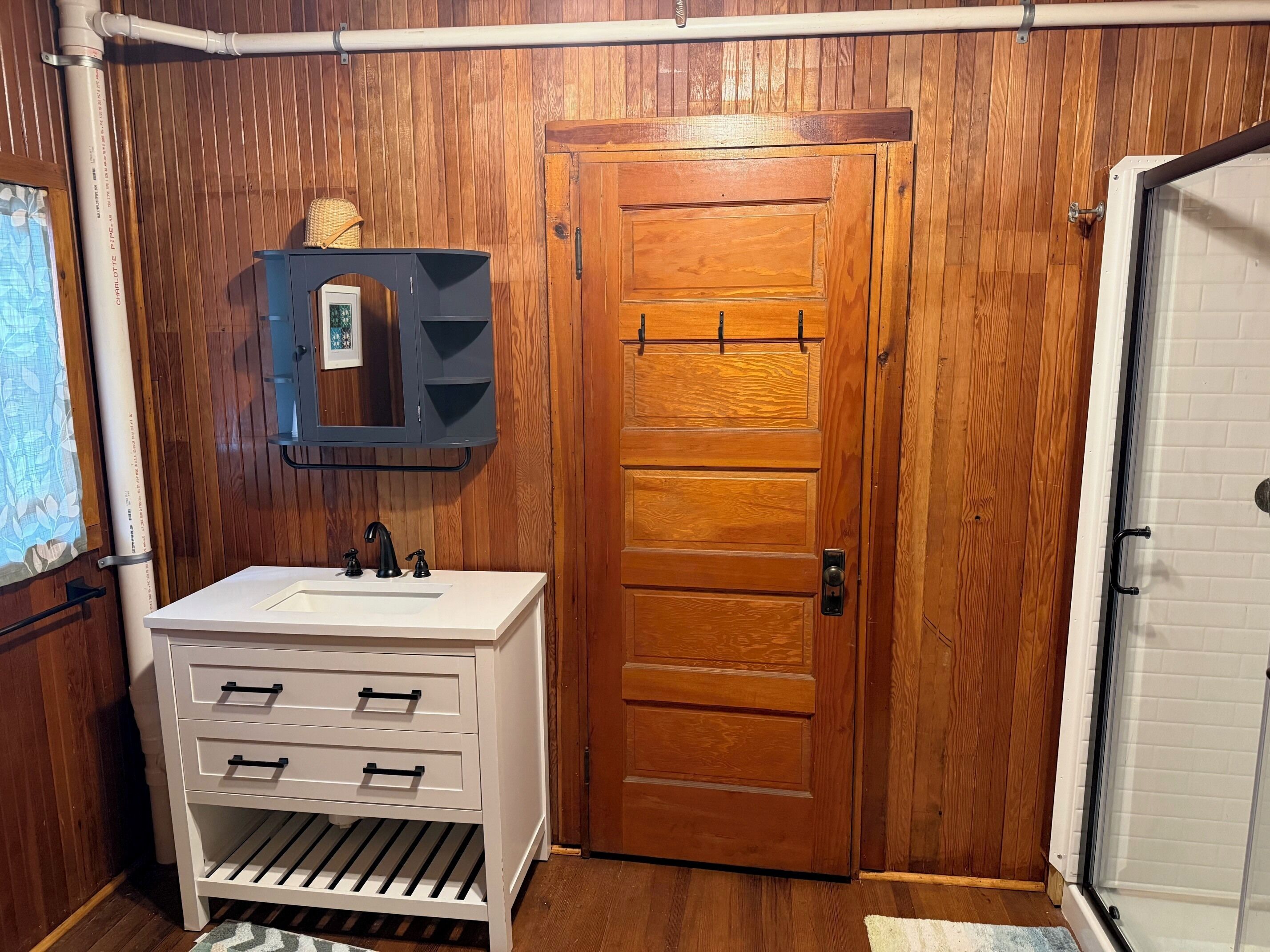 Ein Badezimmer mit Holztür, Waschbecken in Weiß mit schwarzen Armaturen, Handtuchhalter und Duschbereich.