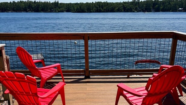 A wooden deck with red chairs facing a body of water and a forested shoreline.