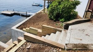 A dock extending into a lake, a wooden staircase leading down to the water, and a small wooden structure with red chairs.