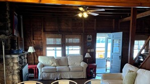 A wooden cabin interior with a dining table, two sofas, a fireplace, and a ceiling fan.