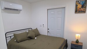 En säng för en person med en metallram, ett sängbord med en lampa, en dörr och en luftkonditioneringsanläggning.