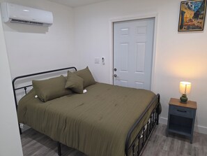 A single bed with a metal frame, a bedside table with a lamp, a door, and an air conditioning unit. - Cozy, quiet guest house with AC in vibrant Hollywood. (Hollywood)