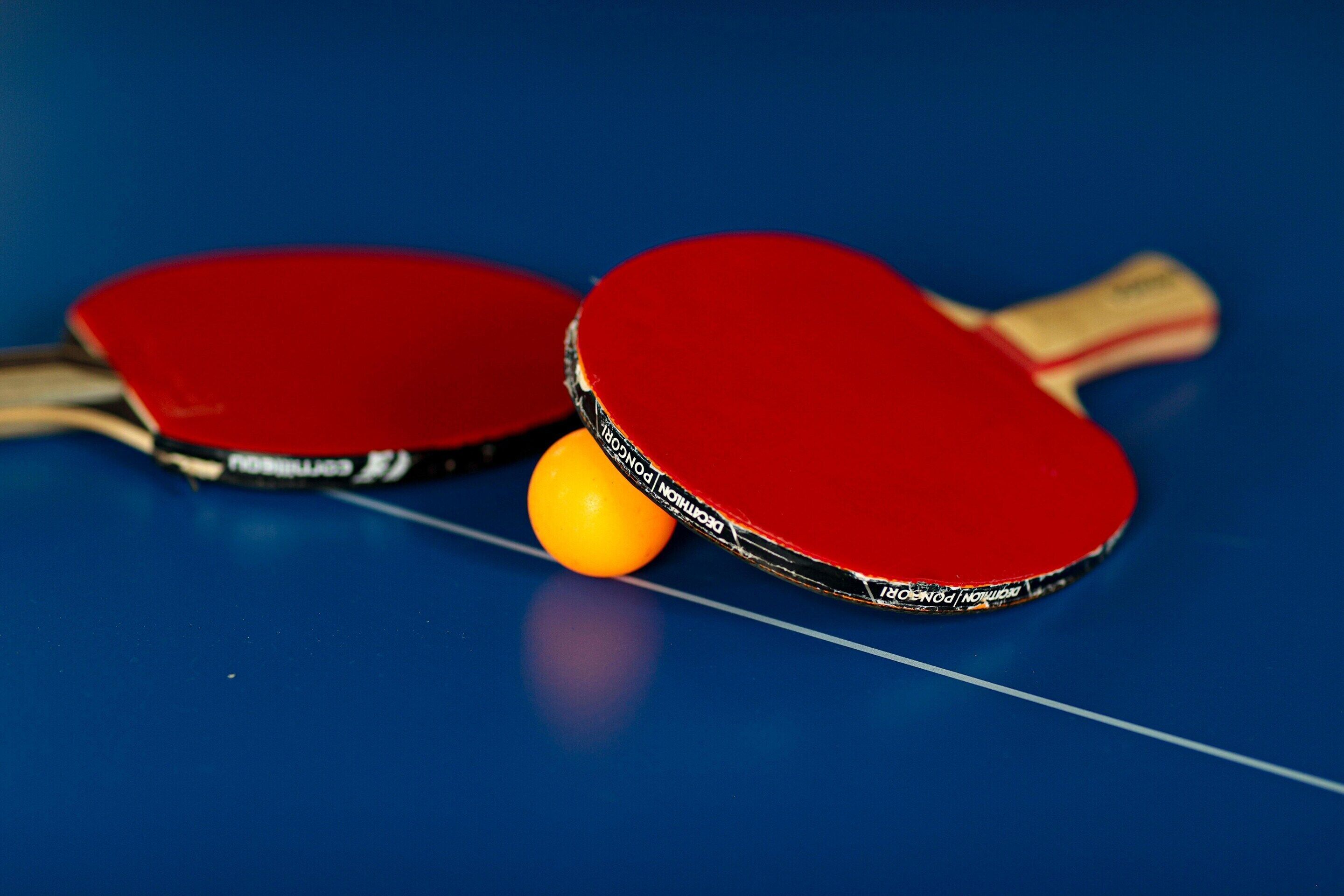 Twee rode tafeltennisrackets en een gele bal op een blauwe tafel.