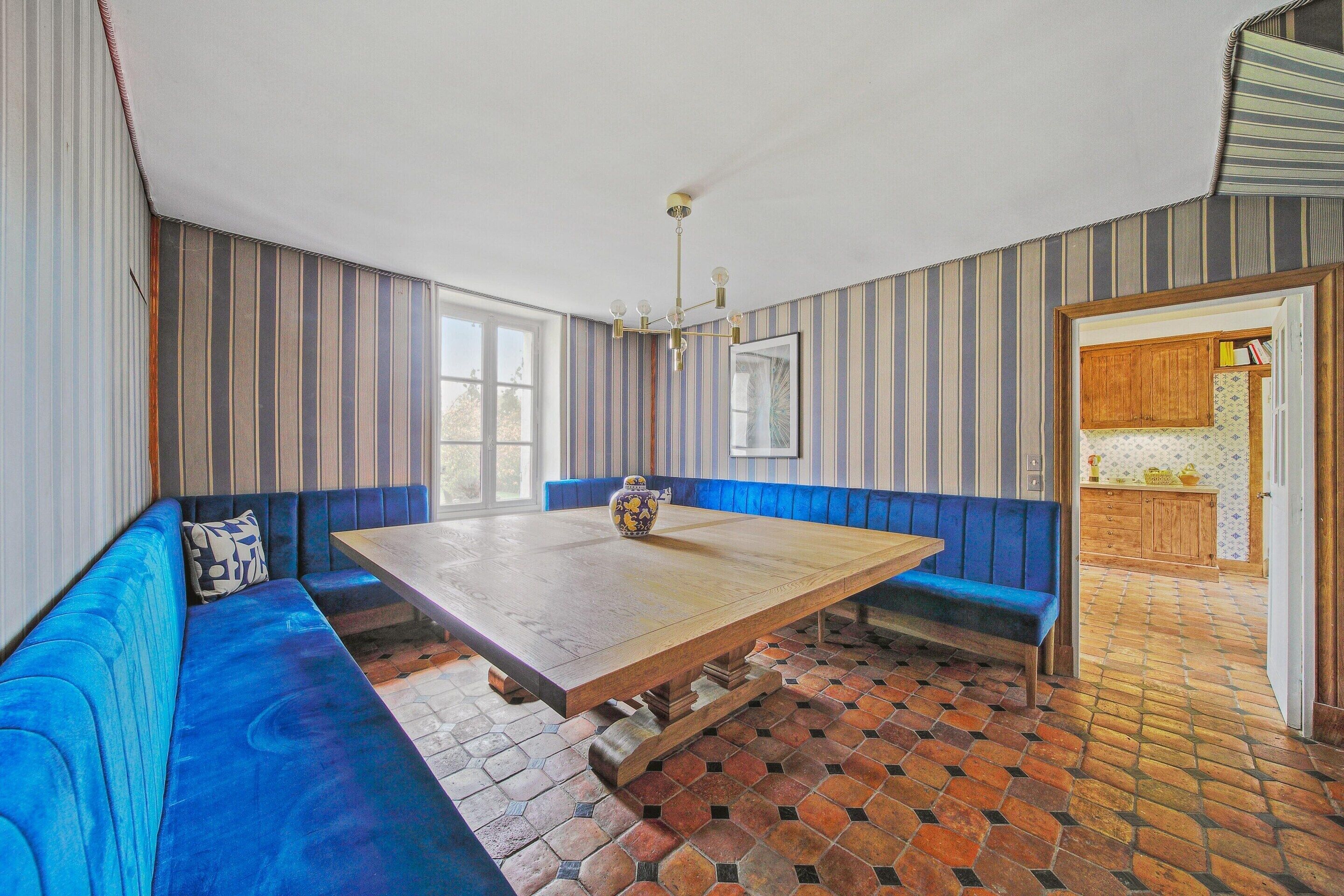 Een eethoek met een lange tafel, blauwe zachte zitplaatsen, een kroonluchter en een keuken die door een open deur zichtbaar is.
