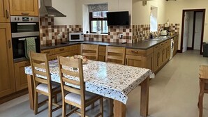 Une cuisine avec des meubles en bois, un îlot central, une table à manger avec des chaises, un four encastré et un téléviseur fixé au mur.