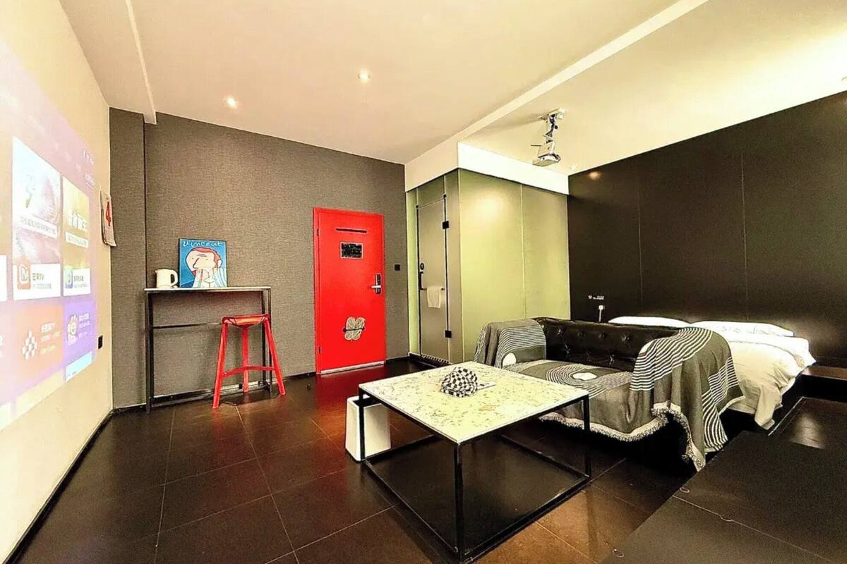 A modern hotel room with a bed, a small table, a red door, and a projector screen on the wall.