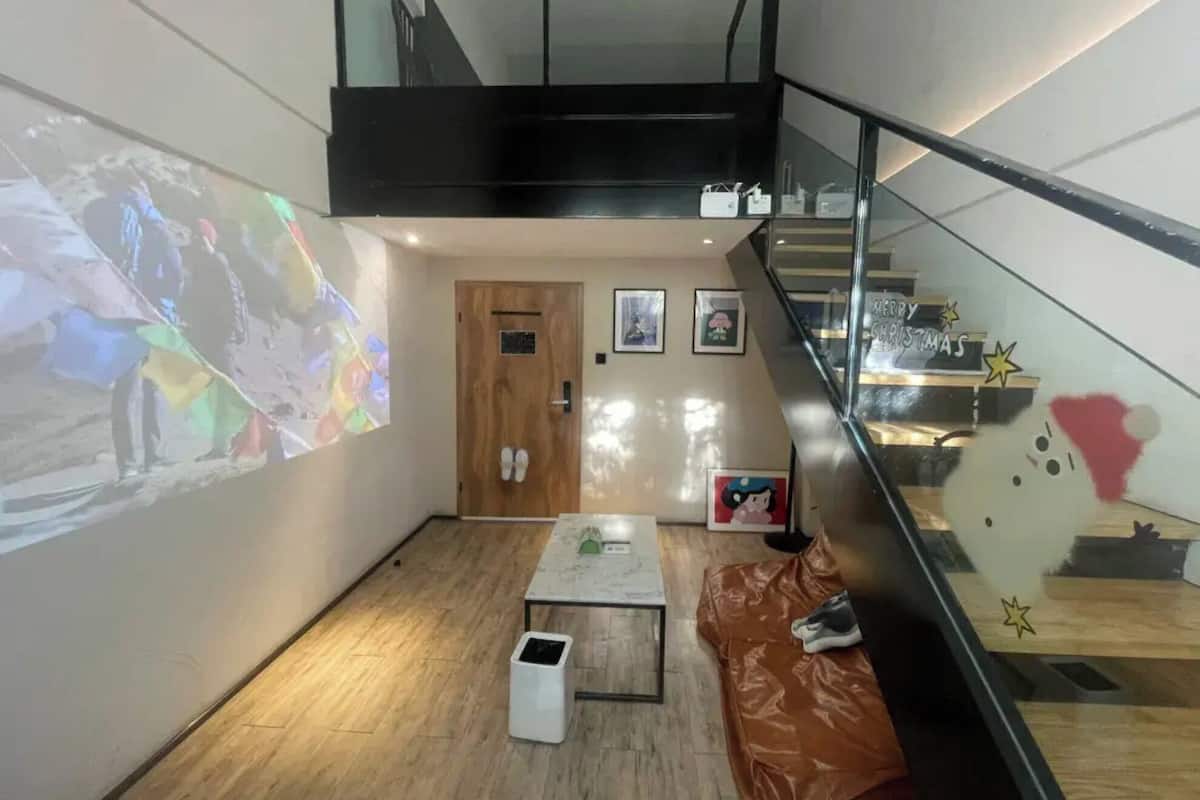 A hotel lobby with a wooden floor, a staircase, a door with a pair of shoes hanging on it, and a small table with a trash bin.