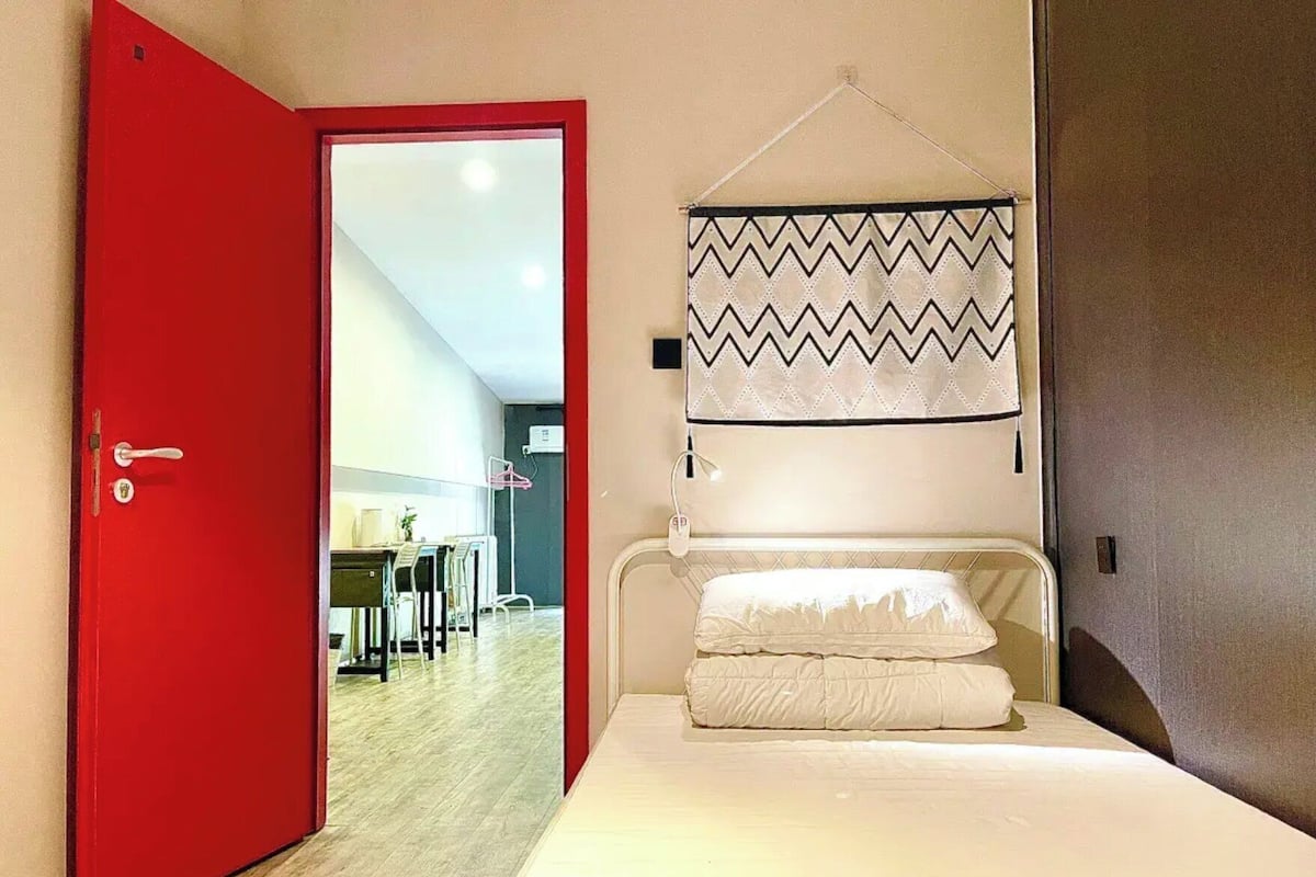 A hotel room with a red door, a bed with white bedding, a hanging patterned curtain, and a view of a dining area through the door.