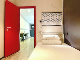 Una habitación de hotel con una puerta roja, una cama con ropa blanca, una cortina colgante con diseño y vistas a un comedor a través de la puerta.