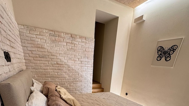 A bedroom with a brick wall, a bed with a quilt, a bedside table with a lamp, and a small rug. - Downtown River Superior New Flat (Lisbon)
