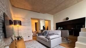 A modern living room with a grey sofa, a wooden side table, a floor lamp, and a brick accent wall. - Downtown River Superior New Flat (Lisbon)