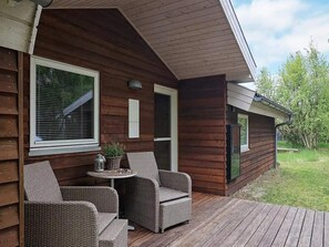 En stuga av trä med en veranda, flätade stolar, ett litet bord och en krukväxt.