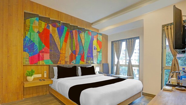 A hotel room with a large bed, a wooden headboard, a colorful abstract painting, a window with curtains, and a television on a stand. - Super Deluxe Room Hotel Kuber Inn (Dhanbad)