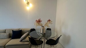 Un salon avec une table à plateau en verre, des chaises noires, un canapé beige, un vase décoratif avec des fleurs et un luminaire fixé au mur.