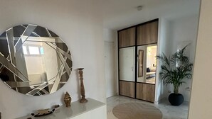 Un intérieur moderne avec un sol en marbre, un grand miroir rond, une console agrémentée d’objets décoratifs, une plante en pot et une vue sur une pièce où se trouve un meuble en bois.