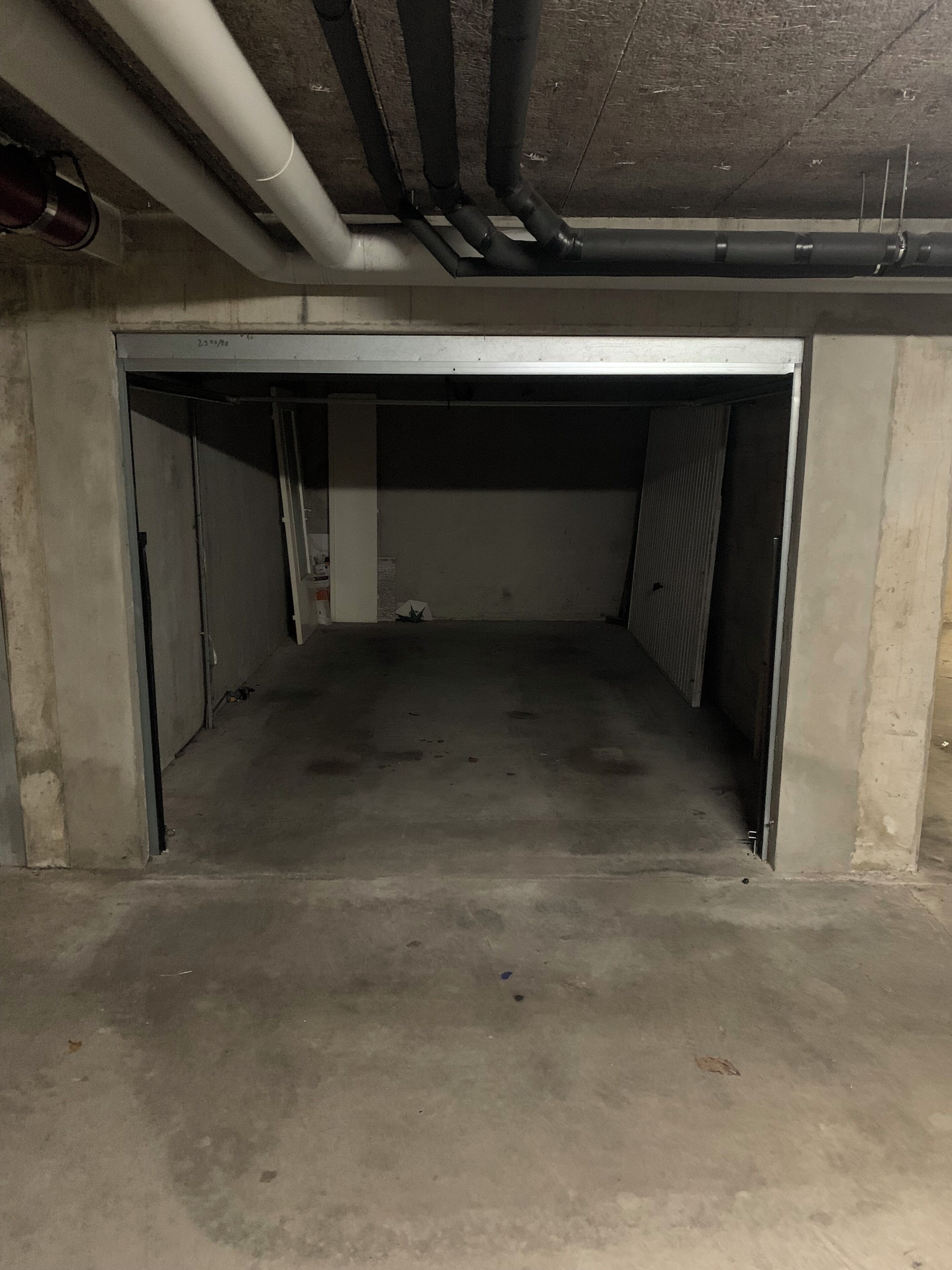 A concrete underground parking garage with a single open door, exposed pipes, and a rough floor.