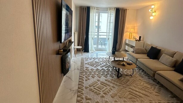 Un salon moderne avec un canapé, une table basse et un téléviseur. On y trouve un tapis à motifs et une vue sur l’extérieur par une fenêtre.