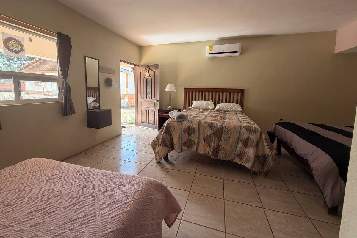 Ein Zimmer mit zwei Betten, einem Deckenventilator, einer Klimaanlage, einem Fenster mit Vorhängen und einer zur Außenseite führenden Tür.