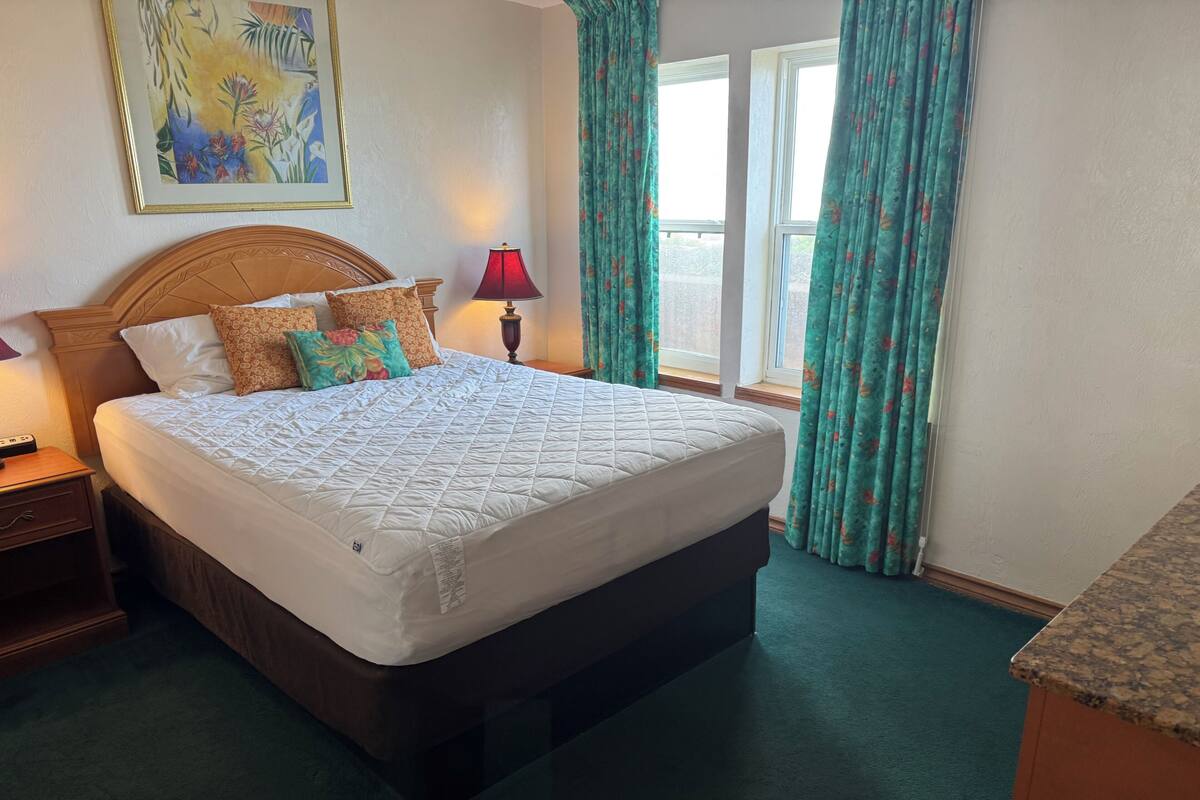 Una habitación de hotel con una cama grande, una mesita de noche, una lámpara, un cuadro en la pared y una ventana con cortinas.