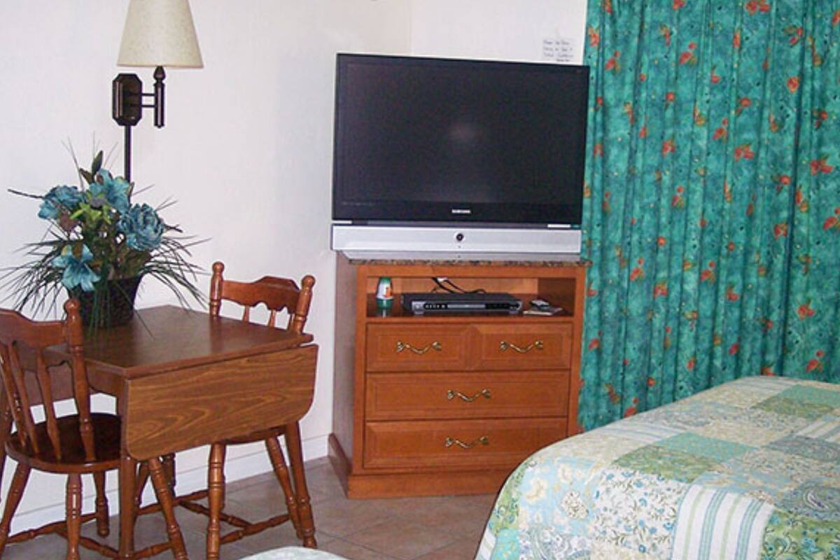Una habitación con una televisión sobre un mueble de madera, una mesa de comedor con sillas y una cama con colcha estampada.