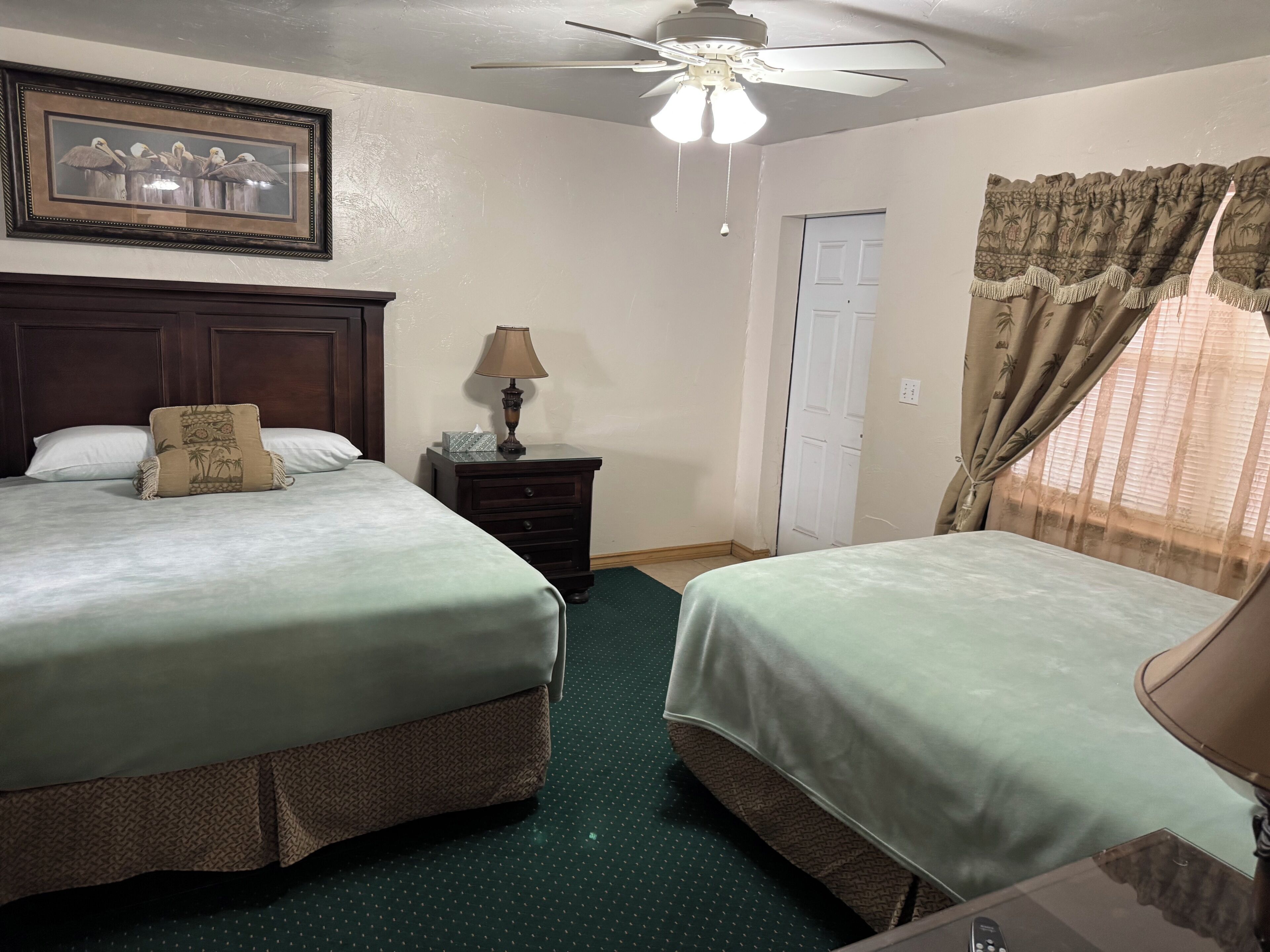 A hotel room with two beds, a ceiling fan, a framed picture on the wall, a nightstand with a lamp, and a window with curtains.