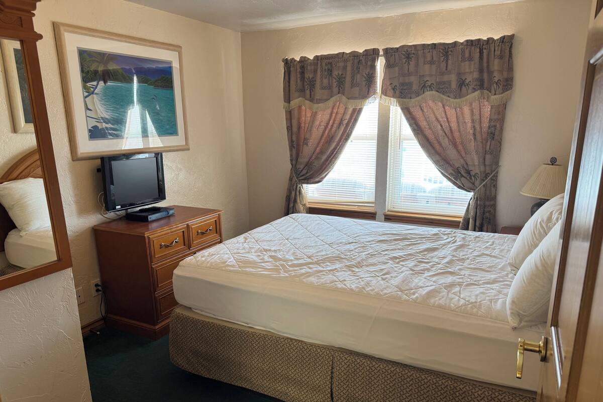 Habitación de hotel con una cama, un televisor sobre una cómoda de madera, una ventana con cortinas y un cuadro en la pared.