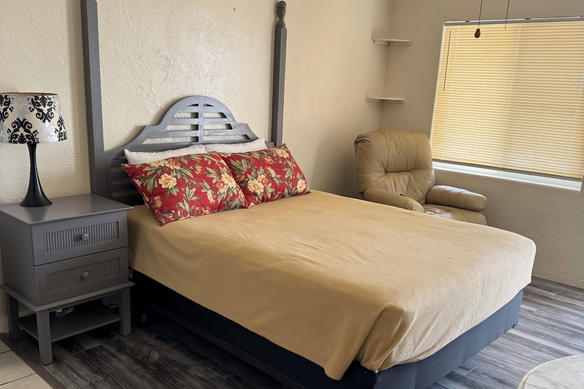 Un dormitorio con cama, mesita de noche, lámpara, silla y ventilador de techo.