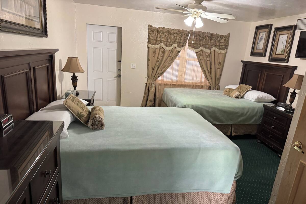 Habitación de hotel con dos camas, un escritorio, un aparador, un ventilador de techo y una ventana con cortinas.