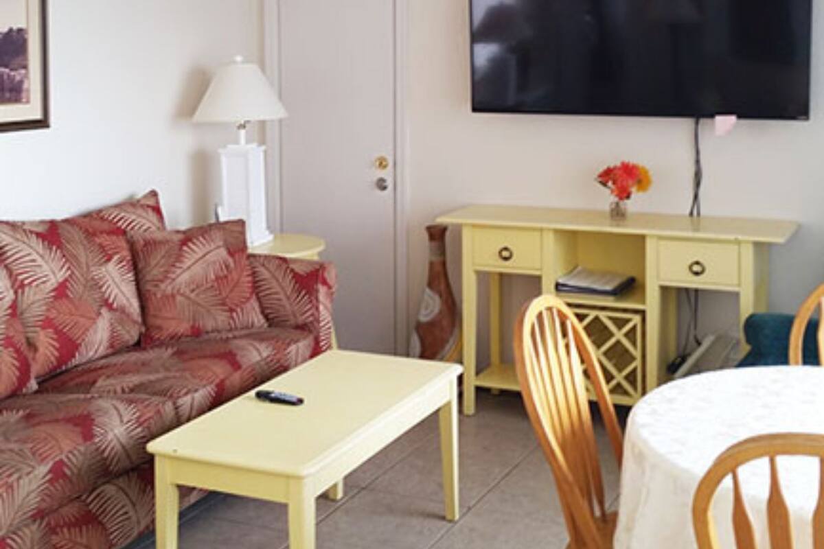 Una habitación con sofá, una mesita, un escritorio con cajones, una silla y un televisor fijado a la pared.