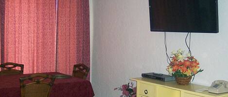 A room with a dining table, chairs, a television mounted on the wall, a small yellow desk with a telephone and a basket, and a potted plant.