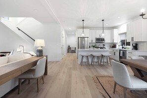 Eine moderne Küche mit weißen Schränken, einer zentralen Kochinsel mit Hockern, einem Essbereich mit langem Tisch und Stühlen sowie einer Treppe, die in ein Obergeschoss führt.