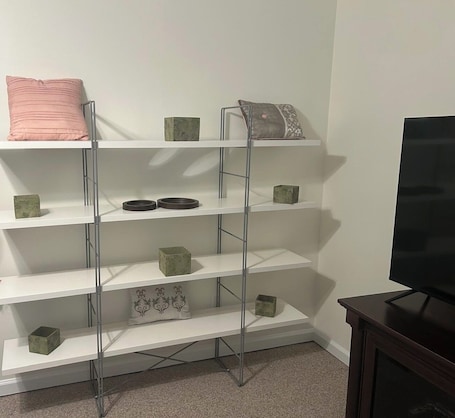 A white metal shelving unit with green and pink cushions, a flat-screen TV, and a dark wooden cabinet.. Cozy, clean 2-bedroom getaway in Attleboro